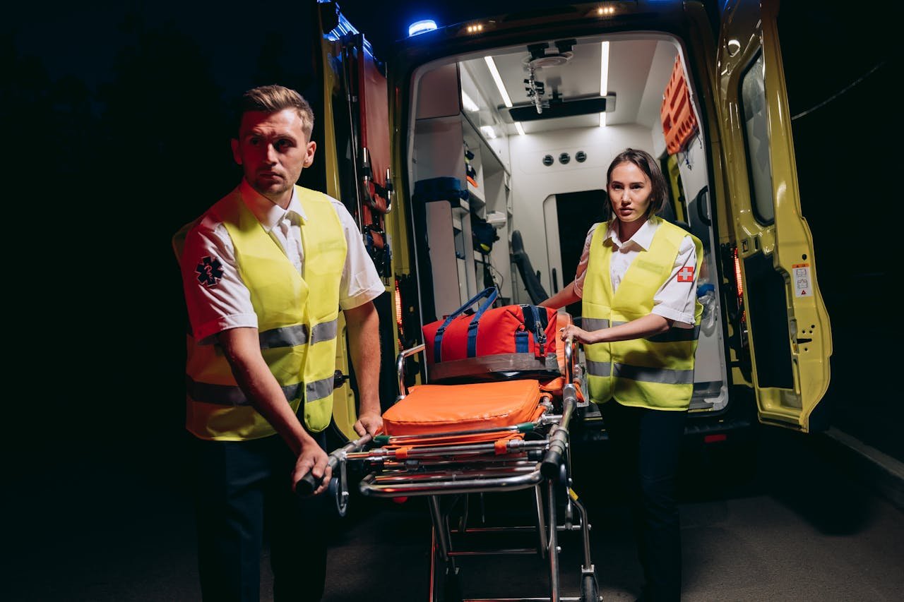 Emergency responders in high-visibility vests prepare a stretcher by an ambulance at night.